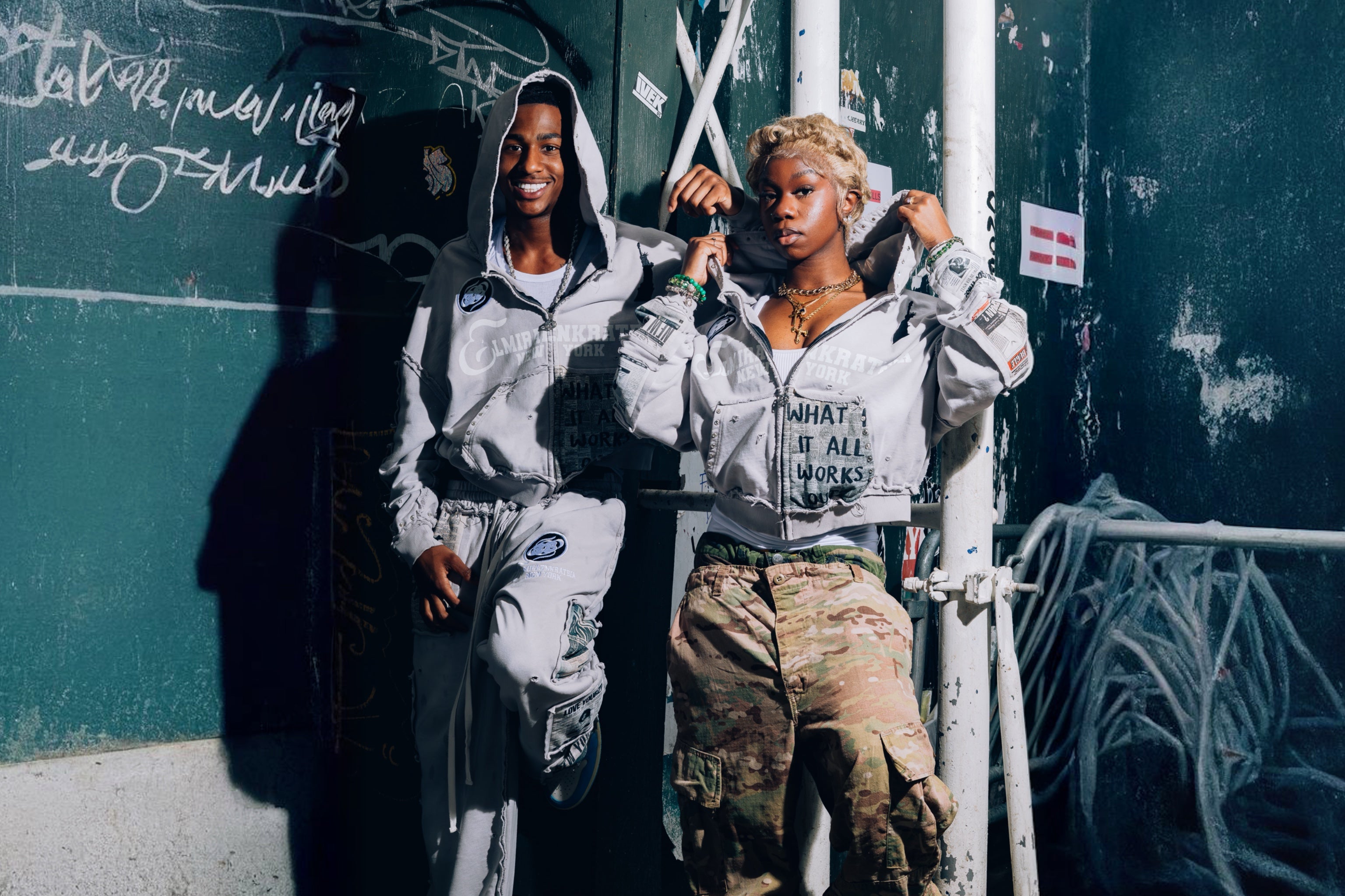 Two people standing against a graffiti-covered wall.